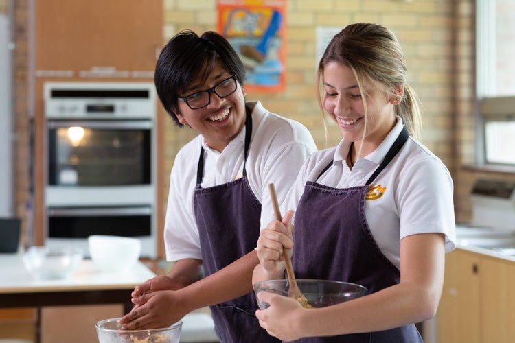 Two students using mixing bowls and stirring ingredients in a kitchen with an oven and other equipment in the background.