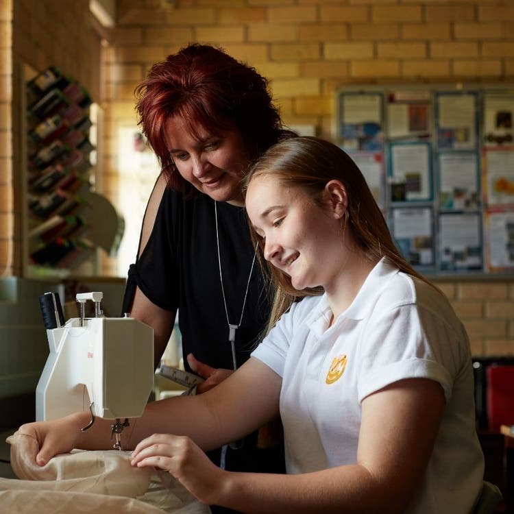 A student in the foreground is using a sewing machine, and a teacher is standing beside her to provide guidance on what to do.