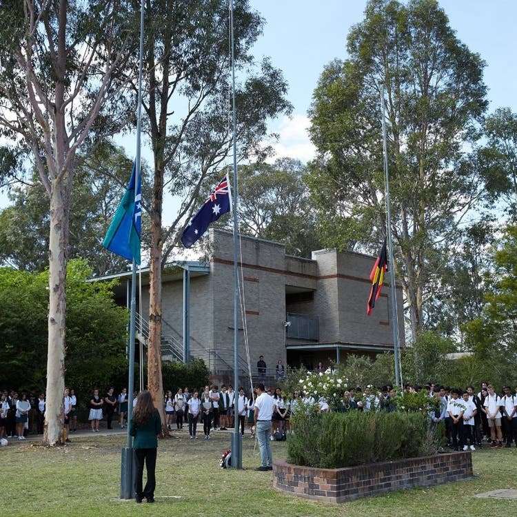 A school assembly around three flag poles, with the Australian flag, the Torres Strait Islander flag and the Aboriginal flag all being at half mast