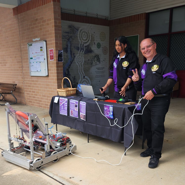A student and a teacher, standing near a display table and operating a robot on wheels using a laptop computer.