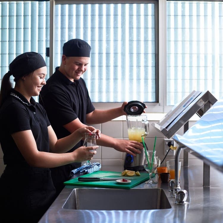 Two students in their hospitality uniforms, using a blender to combine fruit to make drinks which are being prepared in curved glasses with stems