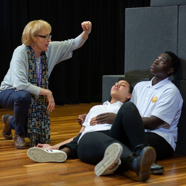 On the left there is a teacher who is giving direction to the two students who are seated on the right of the picture, resting against grey boxes. This photo appears to be of a play which the teacher is providing instruction about.