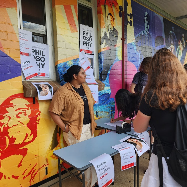 A student standing behind a display table and resting on a wall, which also has posters promoting a student club stuck onto it.