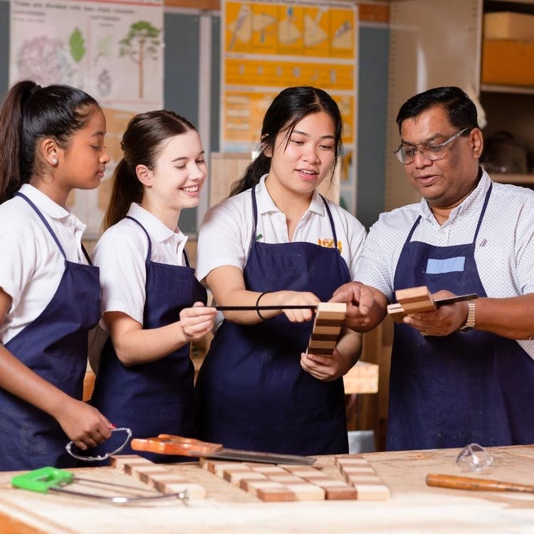 Three students in protective aprons are working in a workshop with timber, being instructed by the class teacher who is on the right of the picture.
