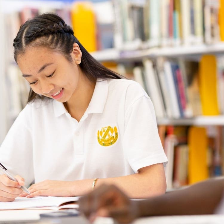 A student sitting at a table in the library, writing in a work booklet.