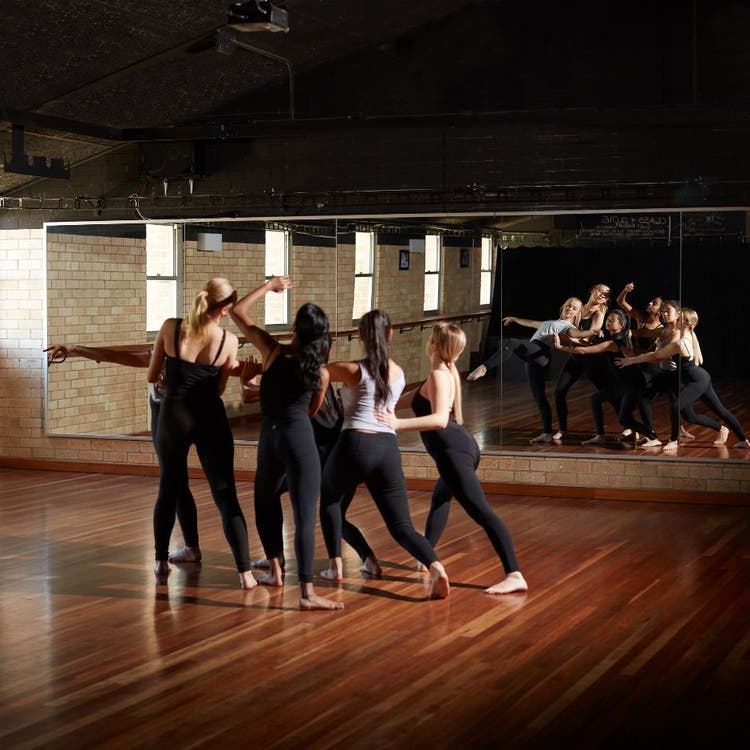 A group of young women dancing as a group in the dance classroom, with their reflection being shown in the mirrored wall in front of them.
