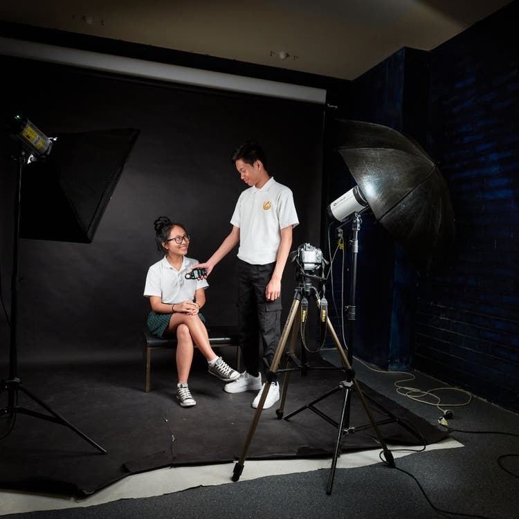Two students are sitting in a photography studio, with a young woman sitting on the chair and a young man is standing near her with a light meter. There are lights focused on the girl as well as a camera on a tripod.