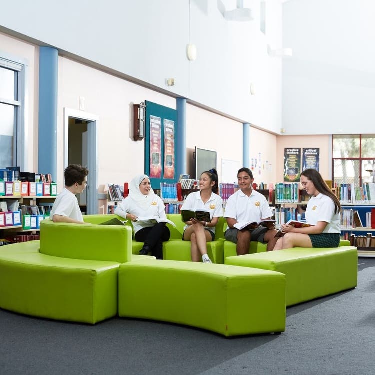 Students sitting on green lounges in the library, talking to each other and looking through books and magazines