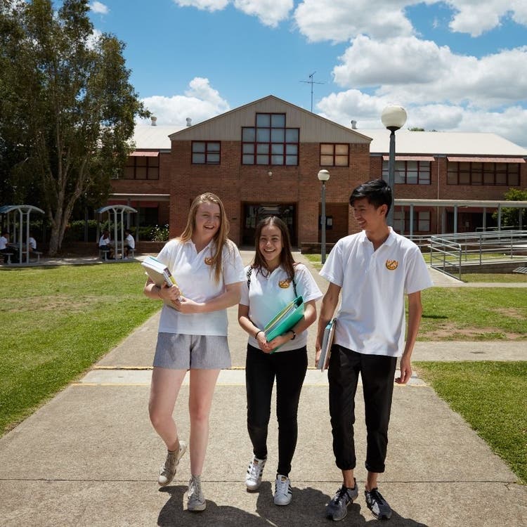 Three students walking across the playground on a concrete path with a large school building in the background.