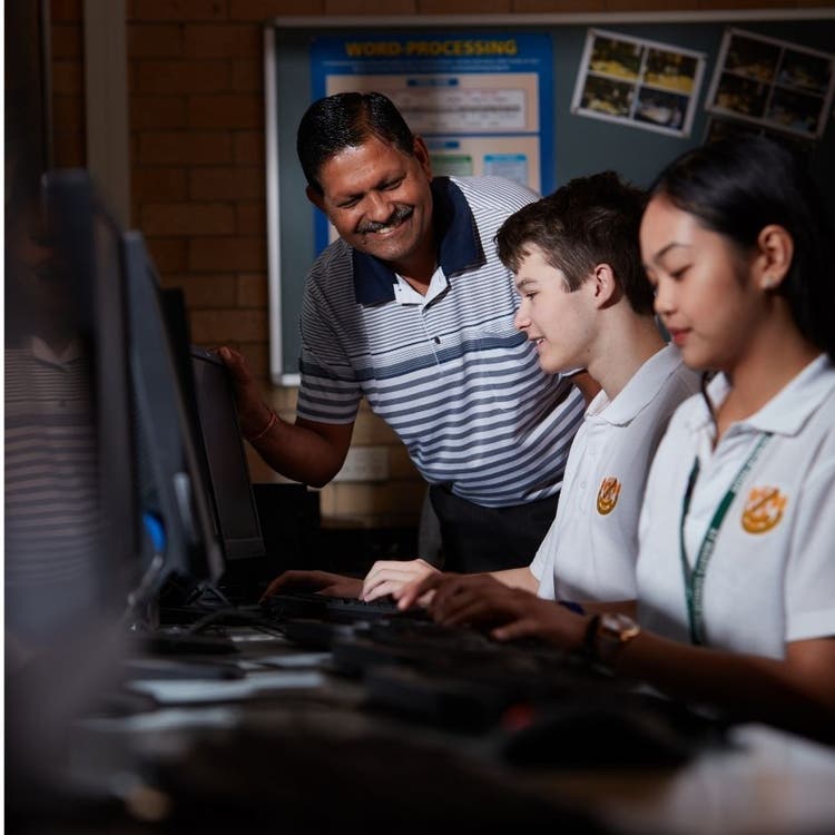 A teacher is in the background, standing and helping two students who are seated in the foreground, typing on computers.