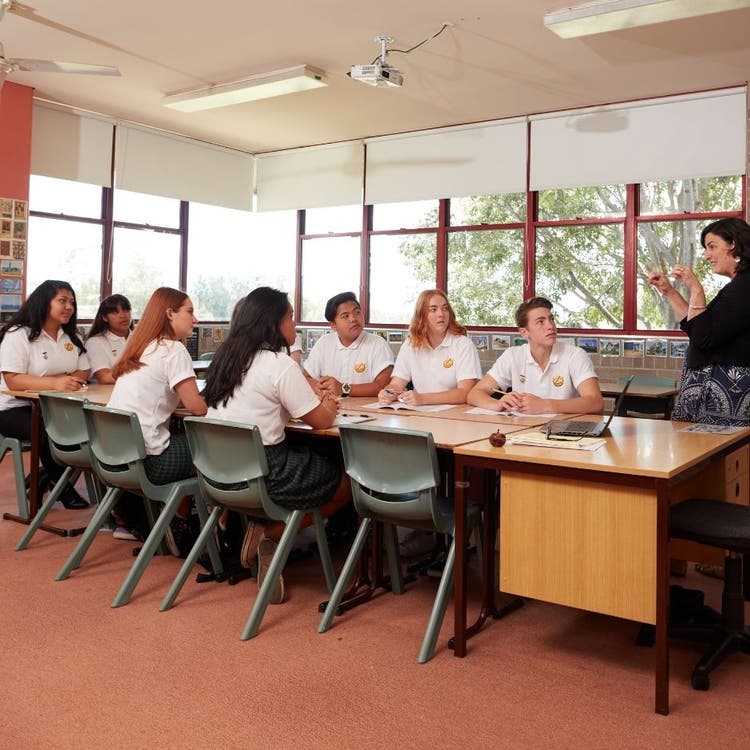 A group of students in a classroom. They are seated in group with their desks facing each other. They are looking forward to a teacher who is shown at the front of the classroom on the right hand side of the picture. She is looking at the students and appears to be explaining something with hand actions.