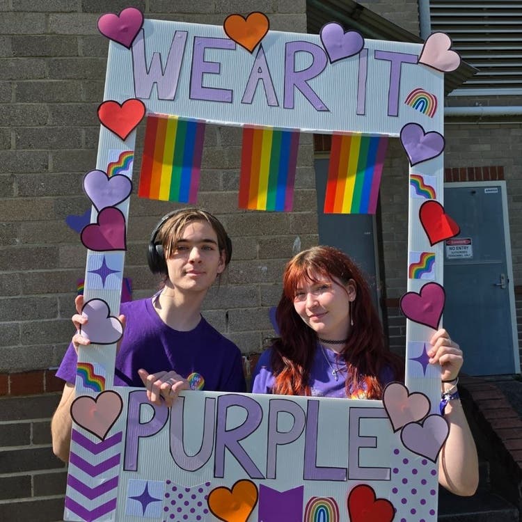Two students dressed in purple posing for a photo while holding a cardboard frame that says WEAR IT PURPLE in the school playground.