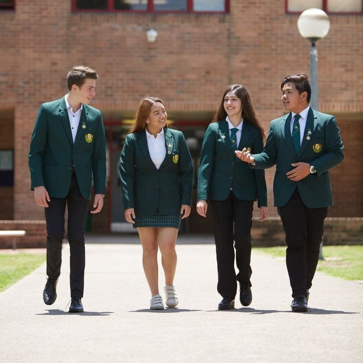 Four students in green blazers and uniform walking on a concrete path in the playground with a large school building in the background.