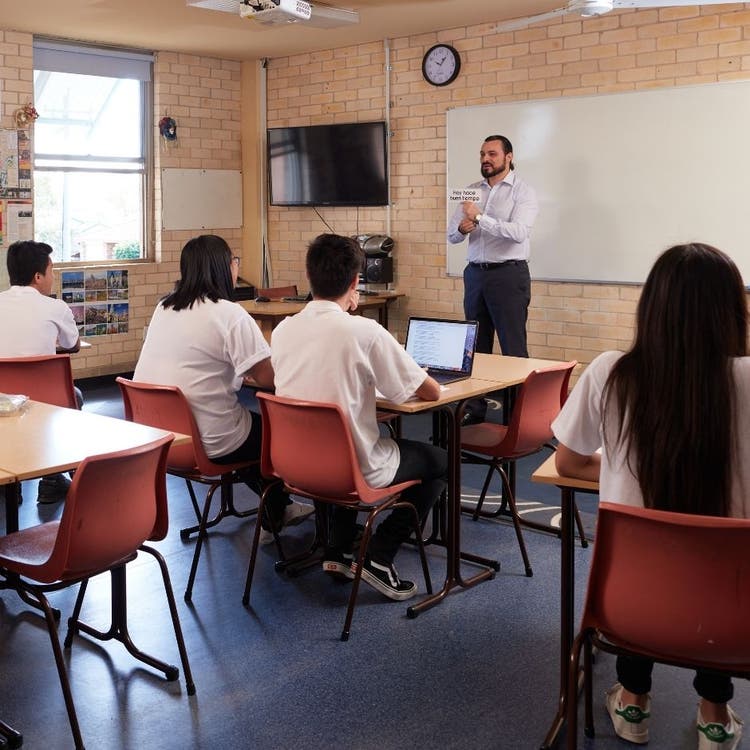 A teacher standing at the front of a classroom providing instruction to students who are sitting at desks. The backs of students can be seen as they look to the front, with the teacher facing the camera.