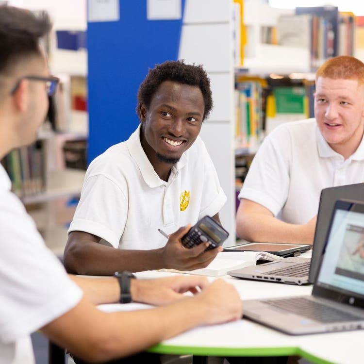 Three students are sitting at desks in the library with shelves of books in the background. In the foreground the three young men have laptops, pens, calculators and books on the tables in front of them and are looking at each other like they are talking to each other.