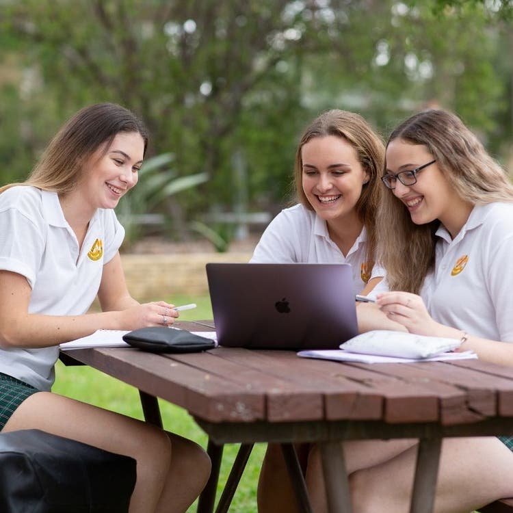 Three students are sitting at a table outdoors and working with paper, pens and pencil cases. The young women are all looking at a laptop screen which is sitting on the table.
