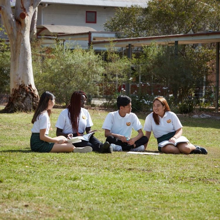 Four students sitting on the grass in the playground. They can be seen from the front but are looking at each other, as if talking to each other and they have some books and equipment with them.