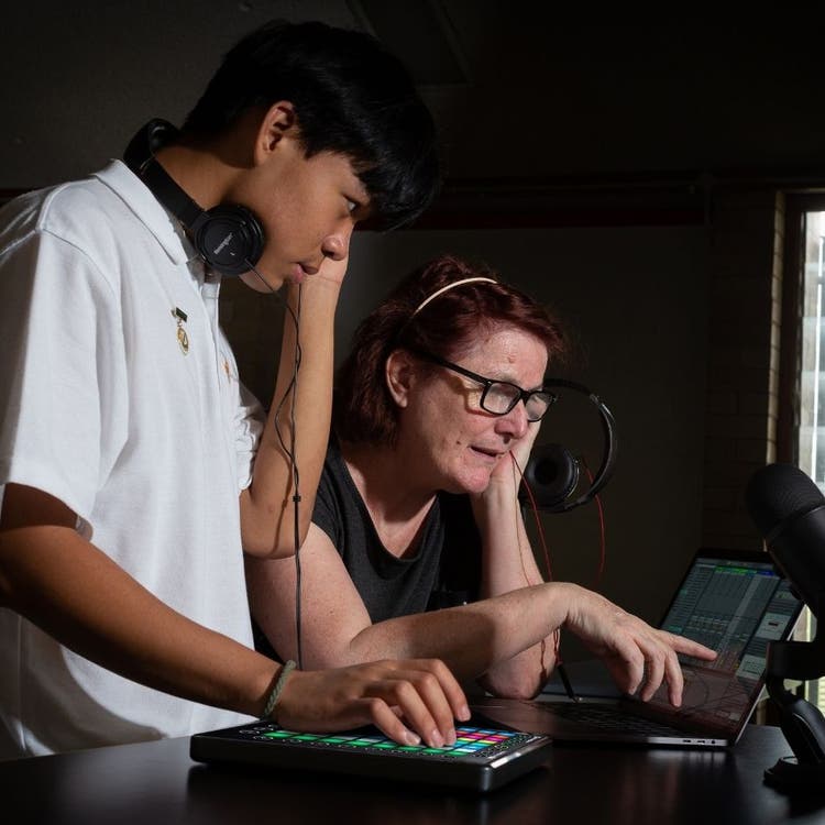 A student is in profile and is standing in the foreground. He has headphones around his neck and is looking at a computer screen while using a touch pad with his right hand. Directly behind him is his teacher, also in profile and she is holding headphones to her left ear and is pointing at the computer screen that the student is looking at.