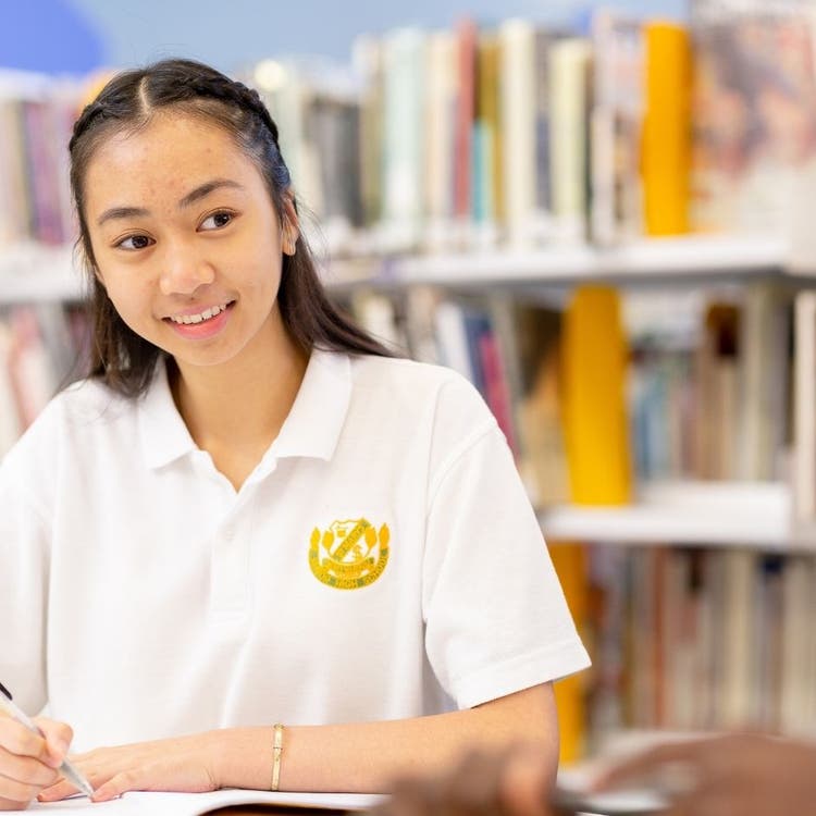 A student is sitting in the foreground, looking forward and writing in a book. In the background there are shelves of books.