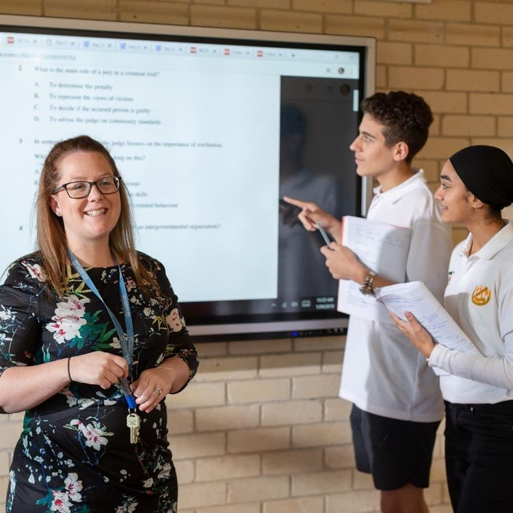 Two students are standing on the right in the background, focused on work on a big display screen on the wall at the back of the picture. In the foreground on the left of the picture, the teacher is directly looking at the camera.