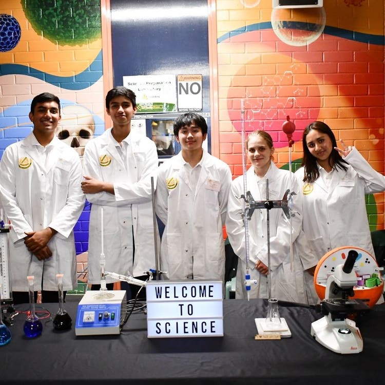Five students in lab coats standing behind a display table which has a range of science equipment on it as well as a sign that says WELCOME TO SCIENCE