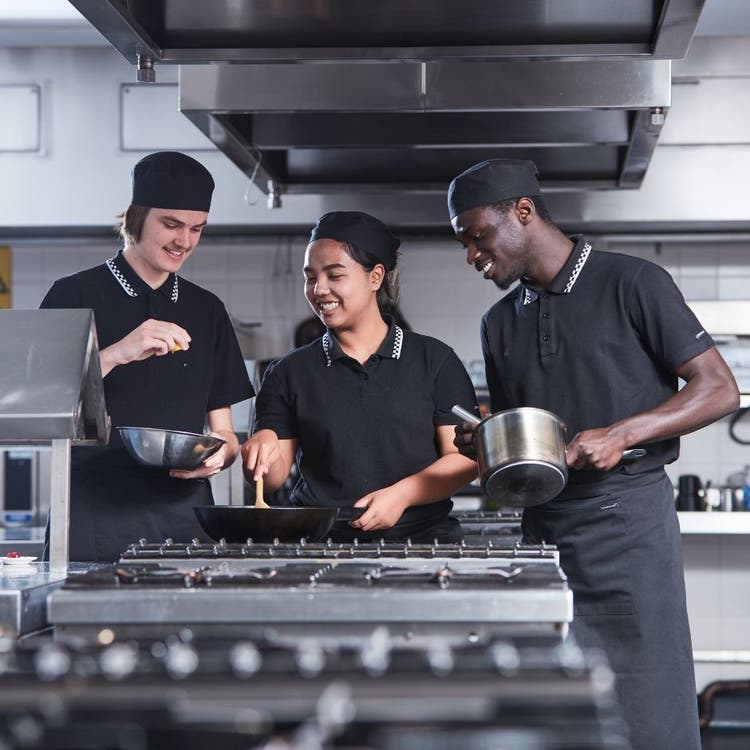 Three students wearing their cooking uniforms, working in a kitchen, standing near a stove top and cooking.