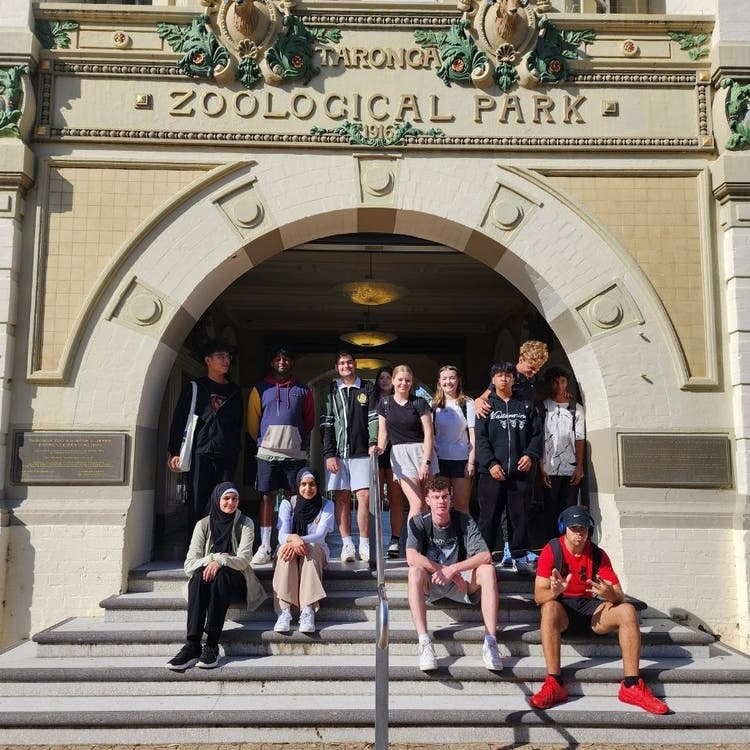 A group of students sitting and standing on concrete stairs with a concrete arch behind them which reads TARONG ZOOLOGICAL PARK on the arch.