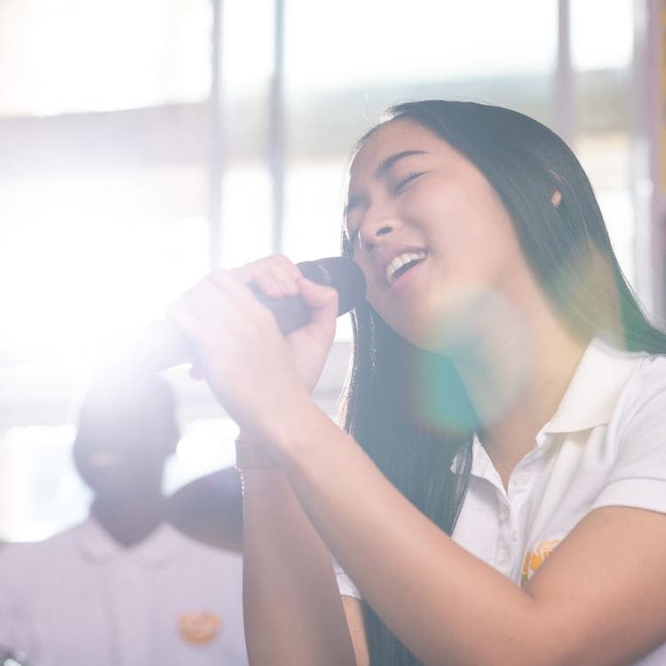 A photo of a young woman singing into a microphone in the music room. She appears in the foreground on the right hand side of the picture. In the background you can see a student sitting and playing the drums.