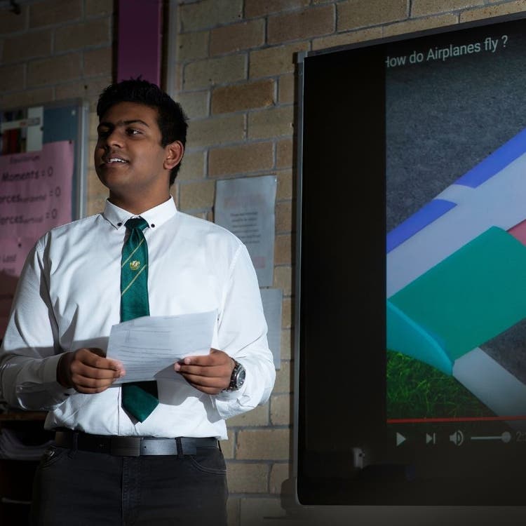 A student standing in front of a screen. He is standing on the left of the photo wearing a white shirt and a green tie, holding a piece of paper. His presentation about planes is shown on the display screen on the wall which is shown on the right of the picture.