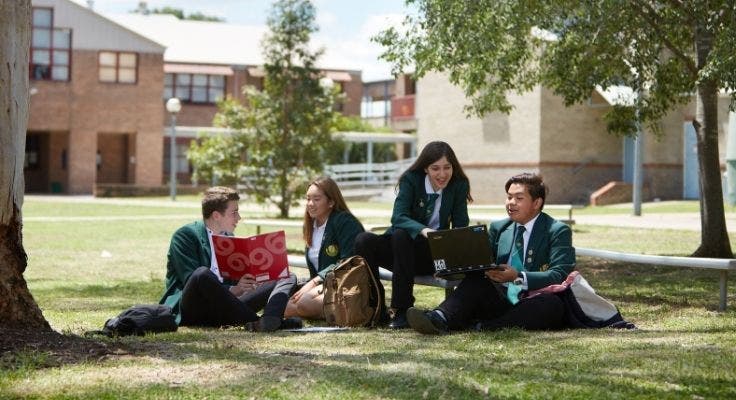 A photo of four students wearing green blazers and dress code, sitting on the grass in the playground under a tree. The students can be seen from the front but are looking at each other, as if speaking to each other about the books and equipment they are holding.